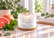 Load image into Gallery viewer, Lit triple-wick Grapefruit &amp; Mint candle in a white vessel on its bamboo lid, beside grapefruit and mint leaves.