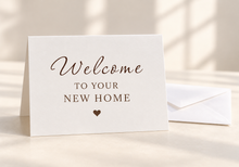 Load image into Gallery viewer, White greeting card reading “Welcome to Your New Home” in espresso brown lettering, with a small heart, beside a white envelope.