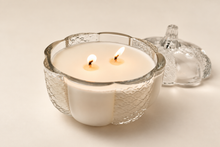 Load image into Gallery viewer, A lit two-wick candle in a textured clear glass pumpkin-shaped vessel, with its matching lid resting behind it.