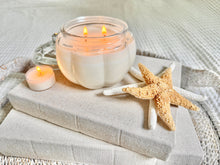Load image into Gallery viewer, Pumpkin-shaped soy candle burning with warm glow on beige books and tray with seashells