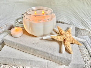 Pumpkin-shaped soy candle burning with warm glow on beige books and tray with seashells